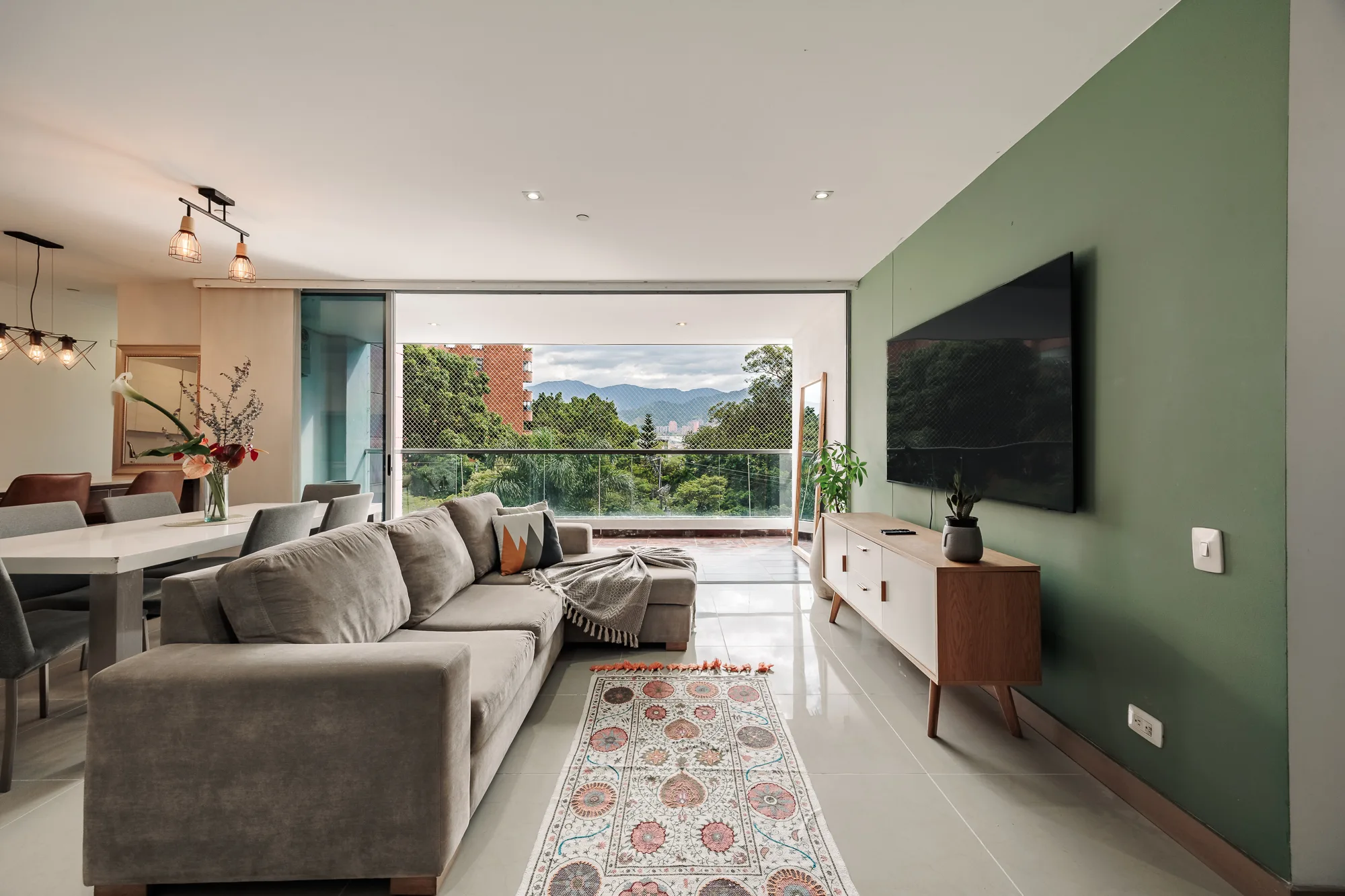 Balcón Verde — bright living room with a sage accent wall, the 80-inch TV and green views over south El Poblado, Medellín